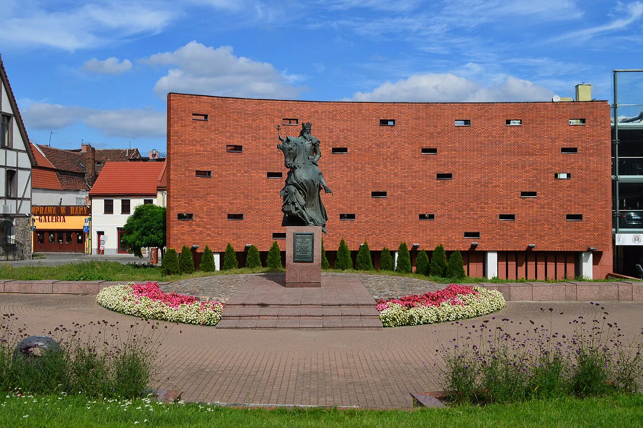 A monument and flowerbeds in front of a red-brick building in Bydgoszcz.